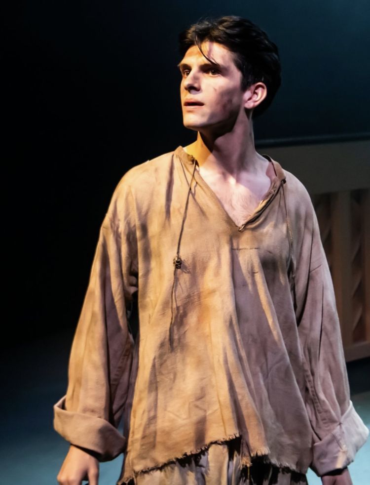 Actor in dirty rag clothing stands onstage looking to his left.